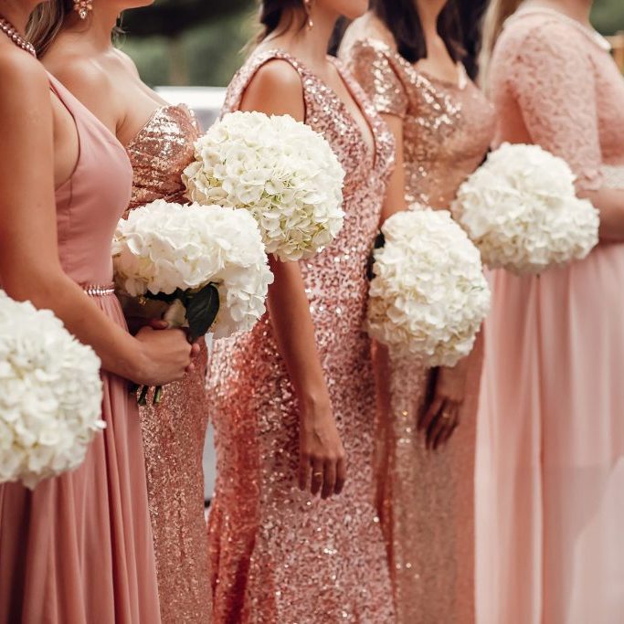 Bridesmaids in pink dresses stand with white flower bouquets in their arms before the arch